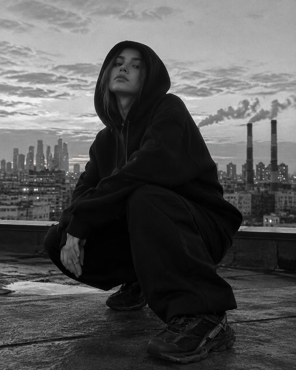 Modelka w czarnej bluzie oversize na dachu z widokiem na industrialne miasto